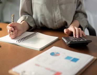 Una persona con una camisa gris escribe en un cuaderno con un bolígrafo mientras usa una calculadora. Sobre el escritorio hay un cuadro impreso con gráficos.