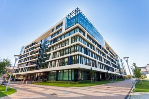 Un moderno edificio de oficinas de varios pisos con exterior de vidrio y concreto, rodeado por una pasarela peatonal y postes de luz, en un día despejado.